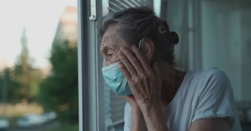 Old woman looking out of window and wearing a mask